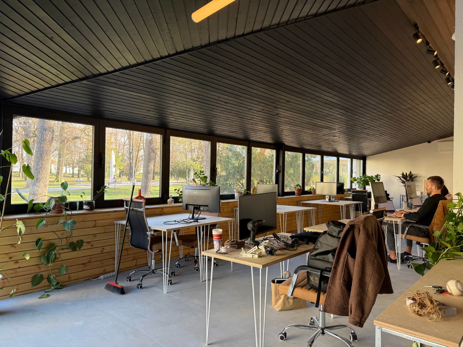 Coworking s pracovnými stolmi pri oknách do parku, jeden člen pracuje za počítačom.