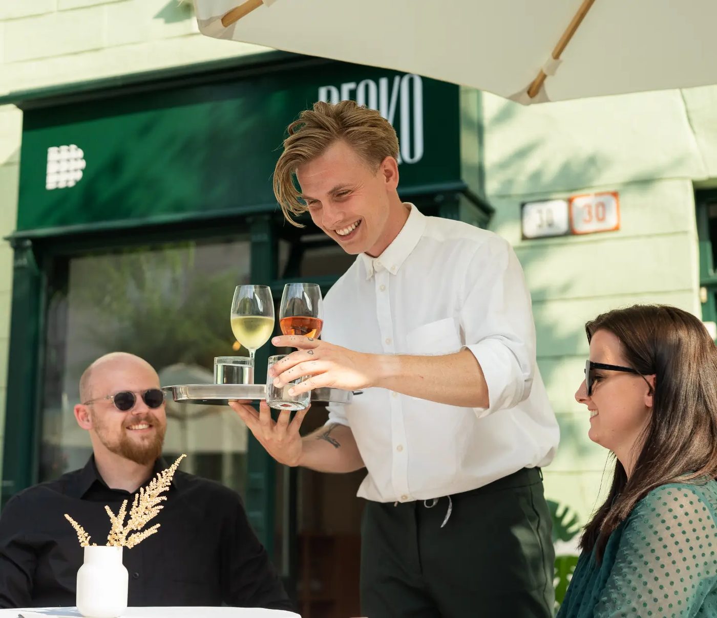Richard v bielej košeli nesie hosťom na terase pred Deckom podnos s pohármi vína.