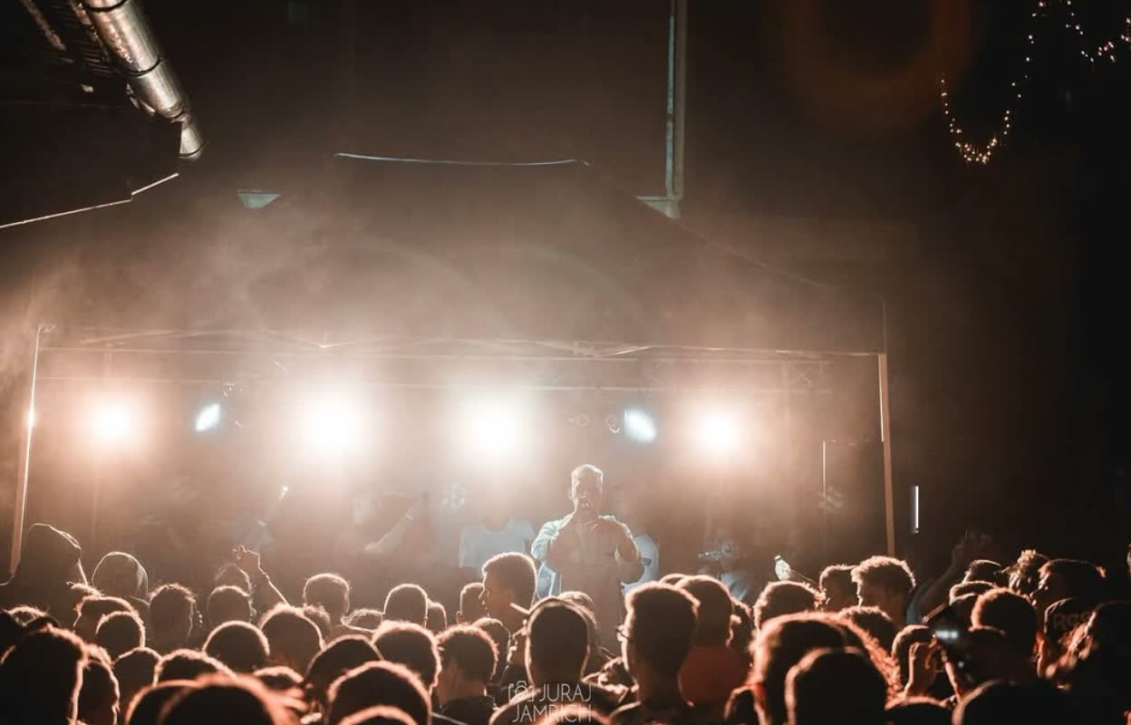 Koncert, raper na pódiu v dyme a svetlách, pred ním dav.
