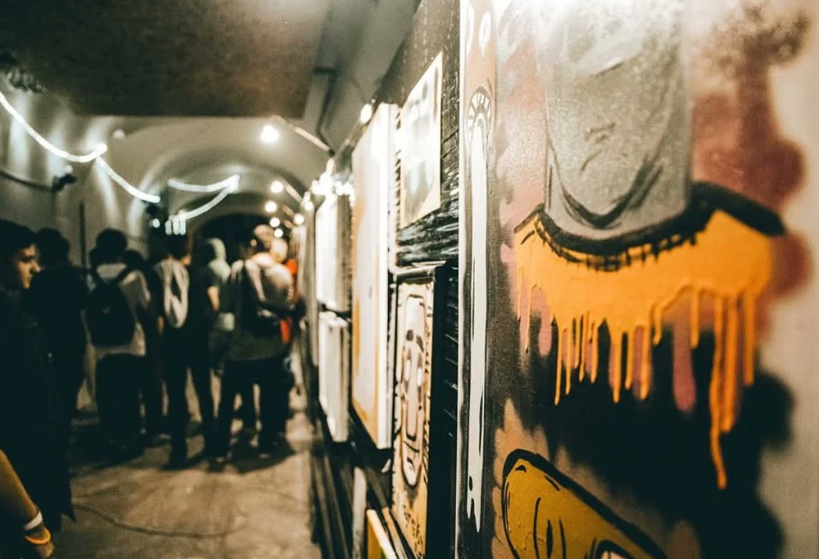 Výstava v klenutej chodbe, obrazy a graffiti na stenách, návštevníci.