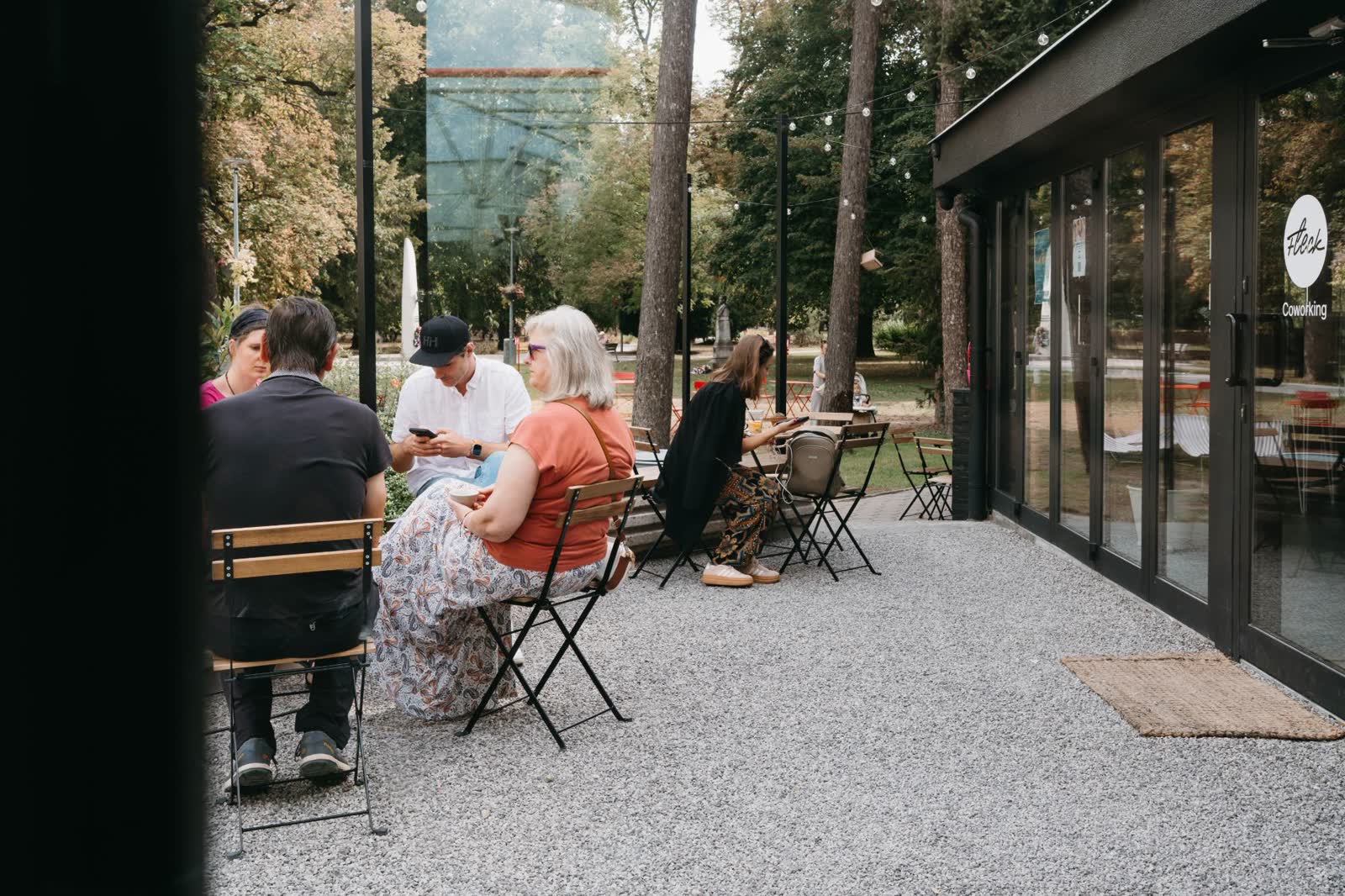 Ľudia sedia na štrkovej terase pri presklenej časti s nápisom Fleck Coworking, v pozadí park.
