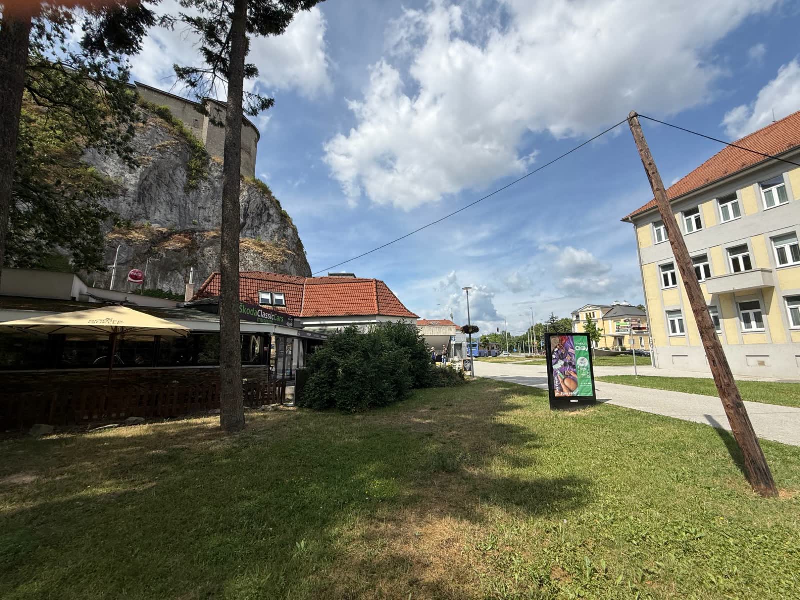 Budova v lete pred kúpou, nápis Škoda Classic Cars múzeum, slnečník a tráva pod hradnou skalou.