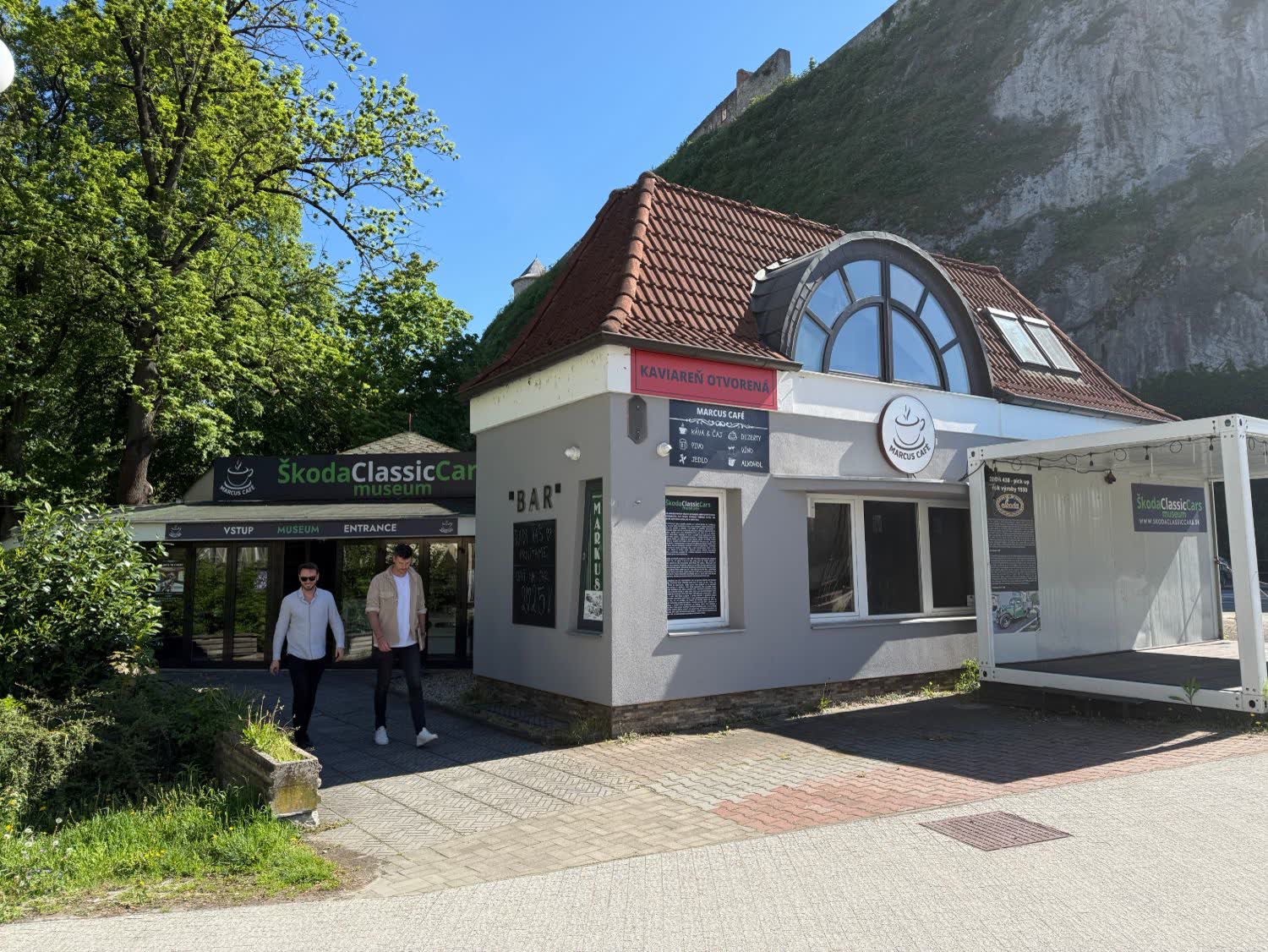Sivá budova s nápisom Kaviareň otvorená a Marcus Café, z dverí vychádzajú dvaja muži.