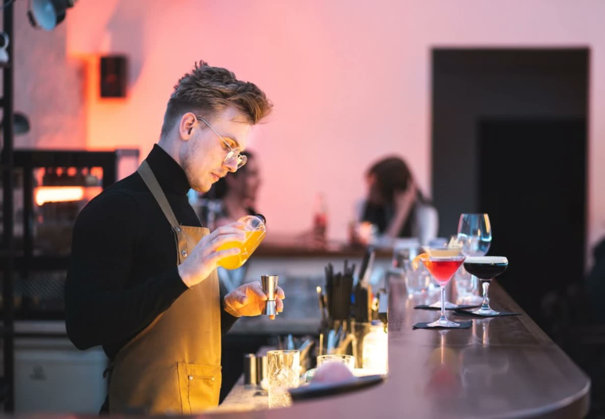 Richard v okuliaroch a zástere mieša drink za barom, na pulte stoja koktaily.
