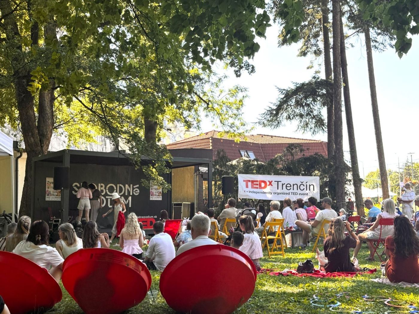 Slobodné pódium v parku počas TEDx Trenčín, ľudia sedia na tráve, na stoličkách a v červených sedačkách pod stromami.