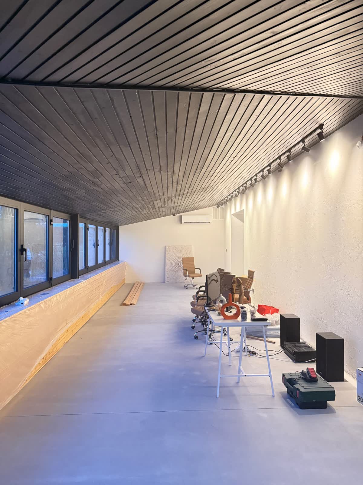 Dlhá hala coworkingu s tmavým lamelovým stropom, fóliou zakrytým parapetom a prvými stoličkami.